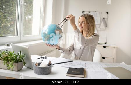 young, beautiful female architect is sitting in her office and is holding a globe and is dreaming of travelling around the world Stock Photo