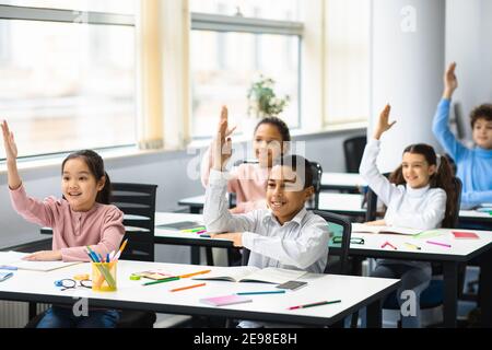 Diverse small schoolkids raising hands at classroom Stock Photo
