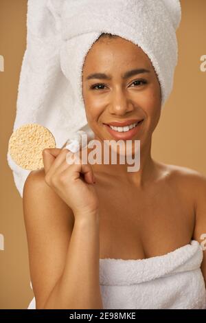 Funny mixed race young woman smiling at camera, holding cleansing face sponge, posing isolated over beige background. Bathroom routine. Youth and skin care concept. Vertical shot Stock Photo