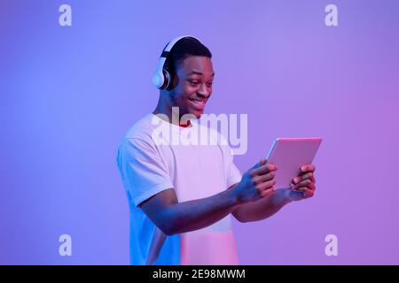 Modern Gadgets. Happy Black Man In Headset Using Digital Tablet, Neon Light Stock Photo