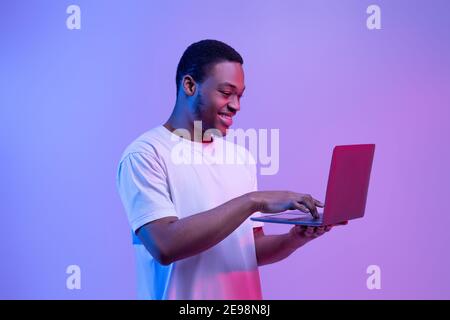 Modern Technologies. Happy Black Man Using Laptop Computer In Neon Light Stock Photo