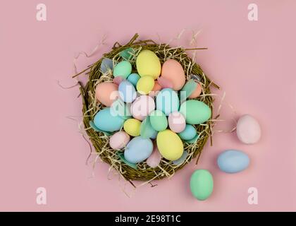 Multi-colored Easter eggs and feathers in pastel colors lie in a nest of branches on a pink background. Copy space. Festive Easter card. Stock Photo
