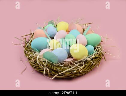 Multi-colored Easter eggs and feathers in pastel colors lie in a nest of branches on a pink background. Copy space. Festive Easter card. Stock Photo