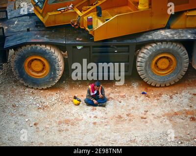 Construction worker resting smoking under a bog truck, Kuala Lumpur Malaysia Stock Photo