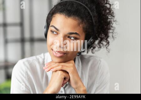 Thoughtful African American millennial woman in a headset is listening to an online webinar, sitting with her hands on a chin, working in the customer Stock Photo