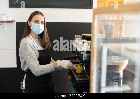 Female barista wearing medical mask preparing coffee take away in a cafe, professional coffee brewing. Small business works during outbreak Stock Photo