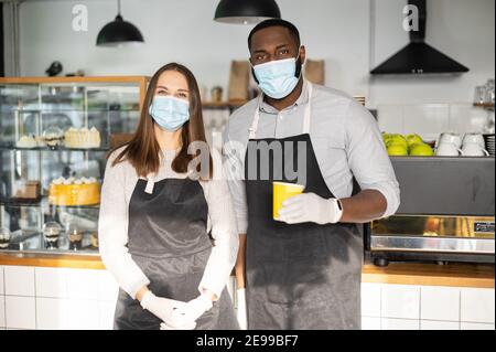 Multiracial waiters in aprons, coffee shop staff wearing medical masks and rubber gloves looks at camera, standing at cozy bakery cafe. Protective Stock Photo