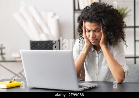 Exhausted millennial African American female office worker touching temples, does not understand the task, suffering from severe headache or migraine Stock Photo