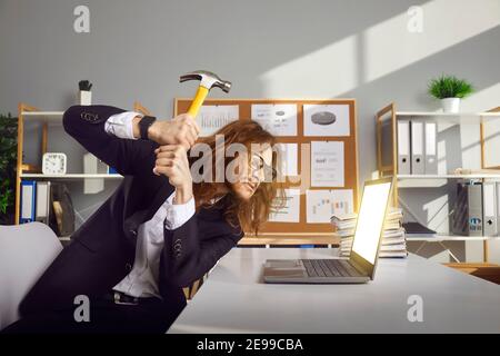 Angry office worker annoyed with computer error or pop up ads smashing laptop with hammer Stock Photo