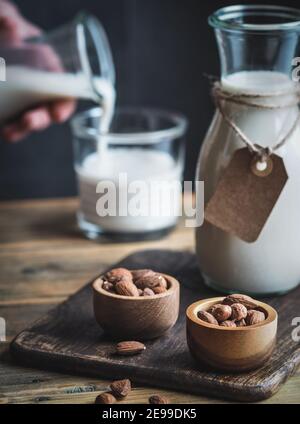 Bottle and glass of pouring almond milk. Empty paper label on bottle. Wooden bowls with almonds on rustic wooden table. Copy space. Stock Photo