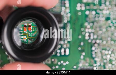 Small electronic components in black loupe on green PCB background. Closeup of magnified red semiconductor diodes, resistor or capacitors in hand lens. Stock Photo
