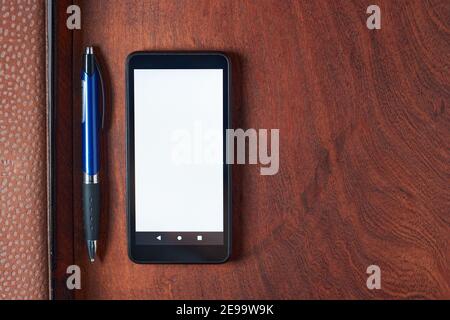 Black mobile phone with blank white screen, pen and notebook on rustic wooden table. Minimalistic top view of office setup, workspace with space for t Stock Photo