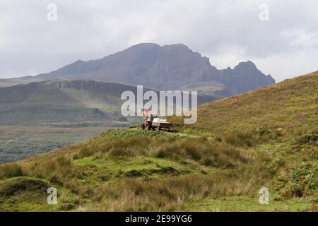 A Red Tractor with a trailer, makes it's lonely way across moorland on the Isle of Skye, Outer Hebrides, Scotland. Bla Bheinn (Blaven) as a backdrop. Stock Photo