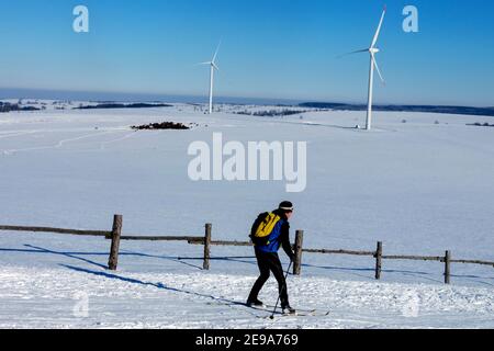 Skier cross country skiing in a snowy landscape, two wind turbines in the background, Czech-German border