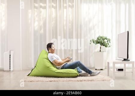 Profile shot of a casual young man sitting on a green bean bag and playing video games with a joystick Stock Photo