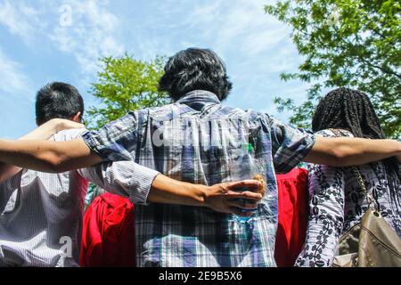 Friends of the graduates - back view of group of multi-ethnic friends with arms around each others shoulders - one holding snacks - talking to graduat Stock Photo