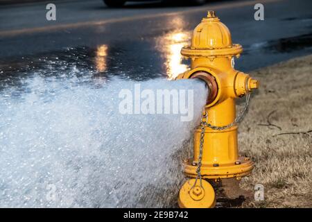 Close-up of yellow fire hydrant gushing water across a street with wet highway and tire from passing car behind Stock Photo