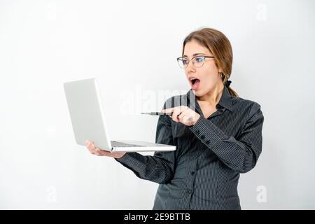 Surprised isolated beautiful young woman posing in study shot holding laptop in hand pointing with pen the screen wearing office business clothes. Cau Stock Photo