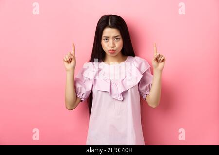 Sad and moody asian girl look from under forehead and sulking, pointing fingers up and complaining, staring jealous or upset, standing against pink Stock Photo