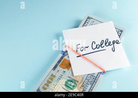 Text written note FOR COLLEGE with dollars cash money in rubber band, on copy space blue background - concept of financial planning to save money for Stock Photo