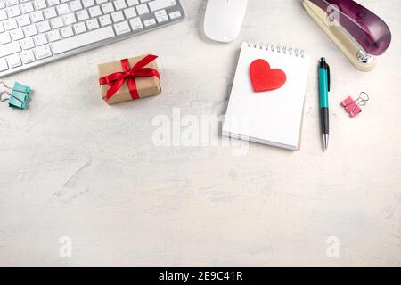 Red heart on notepad near keyboard, pen, mouse, stapler and gift with red ribbon on workplace. Valentine's Day in the office. Top view Stock Photo