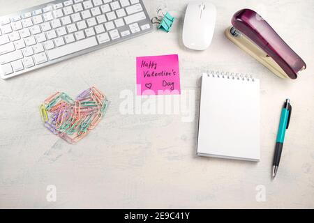 Words Happy Valentine's Day in a note on the desktop with colored paper clips in the shape of a heart and keyboard, pen, notepad and stapler at workpl Stock Photo