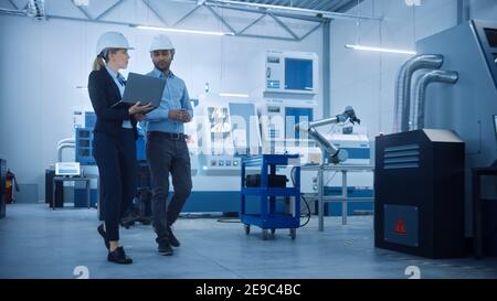Female Chief Engineer and Male Project Manager Walk Through Modern Factory, Talking and Planning Optimization of the Production. Industrial Facility Stock Photo