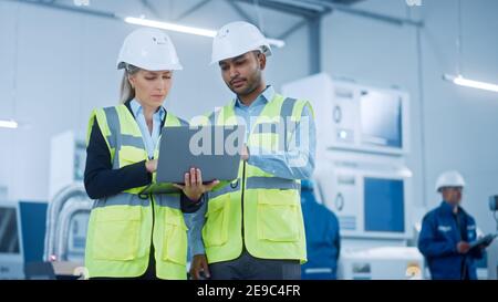 Chief Engineer and Project Manager Wearing Safety Vests and Hard Hats, Use Laptop in Modern Factory, Talking, Optimizing Production Line. Industrial Stock Photo
