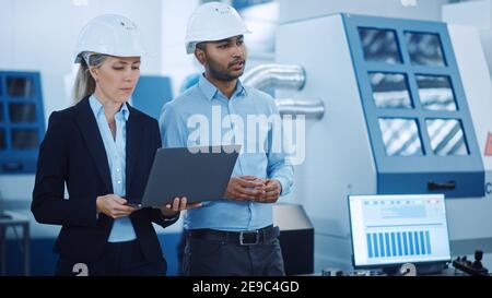 Chief Engineer and Project Manager Wearing Safety Vests and Hard Hats, Use Laptop in Modern Factory, Talking, Optimizing Production Line. Industrial Stock Photo