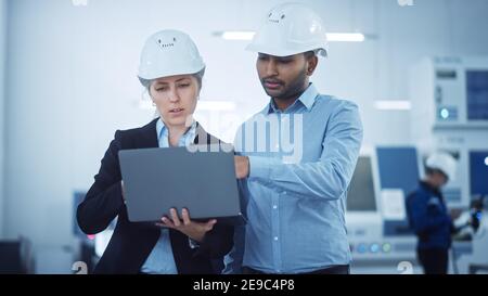 Female Chief Engineer and Male Project Managers Standing in the Middle of Modern Factory, Use Laptop Computer, Plan Production Line Optimization Stock Photo