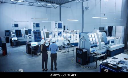 Modern Factory: Two Engineers Use Digital Tablet Computer with Augmented Reality Visualizing Workshop Room Mapping, Floor Layout. Facility with High Stock Photo