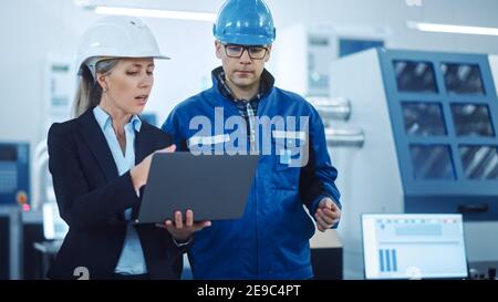Female Manager and Project Engineer Wearing Hardhats Use Laptop in Industrial Factory, Talk, Plan Productivity Optimization. Production Line Workers Stock Photo