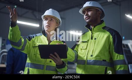 Chief Engineer and Project Manager Wearing Safety Vests and Hard Hats, Use Digital Tablet Computer in Modern Factory, Talking, Optimizing Production Stock Photo