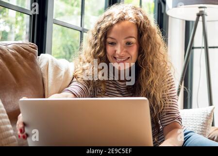 Pretty smiling caucasian woman making video call with laptop computer on the couch in living room at home during day time Stock Photo