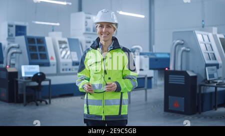 Beautiful Smiling Female Engineer Wearing Safety Vest and Hardhat Holds Safety Goggles. Professional Woman Working in the Modern Manufacturing Factory Stock Photo