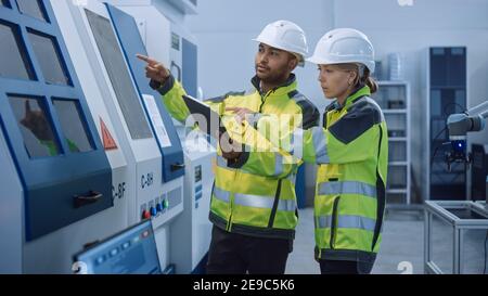 Chief Engineer and Project Manager Wearing Safety Vests and Hard Hats, Use Digital Tablet Controller in Modern Factory, Talking, Optimizing CNC Stock Photo