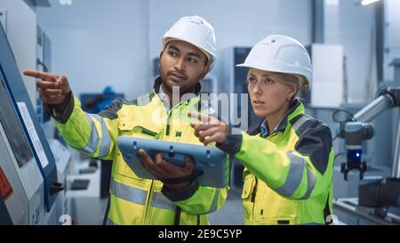 Chief Engineer and Project Manager Wearing Safety Vests and Hard Hats, Use Digital Tablet Controller in Modern Factory, Talking, Optimizing CNC Stock Photo