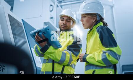 Chief Engineer and Project Manager Wearing Safety Vests and Hard Hats, Use Digital Tablet Controller in Modern Factory, Talking, Programming Machine Stock Photo