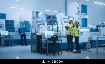 Chief Engineer and Project Manager Wearing Safety Vests and Hard Hats, Use Digital Tablet Controller in Factory, Optimizing CNC Machinery Programming Stock Photo