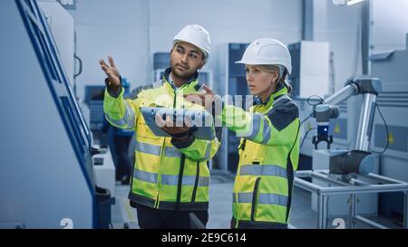 Chief Engineer and Project Manager Wearing Safety Vests and Hard Hats, Use Digital Tablet Controller in Modern Factory, Talking, Optimizing CNC Stock Photo