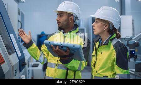 Chief Engineer and Project Manager Wearing Safety Vests and Hard Hats, Use Digital Tablet Controller in Modern Factory, Talking, Optimizing CNC Stock Photo