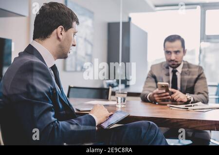 Two businessmen using smart phones and touchpad at meeting. Stock Photo