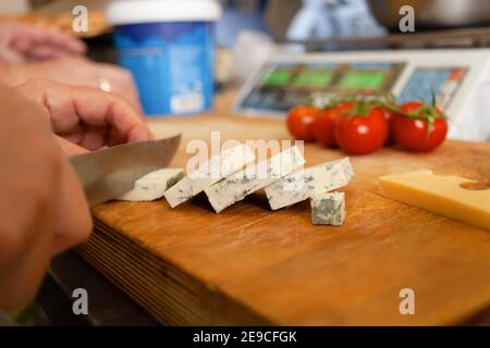 A woman cuts cheese with mold on a wooden board. In the background are scales and tomatoes. Stock Photo