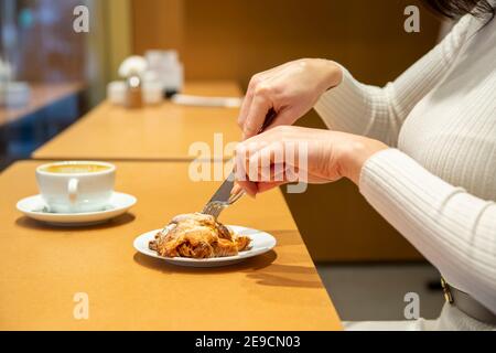 woman cuts croissant and drinks coffee at a table in a cafe. no face Stock Photo