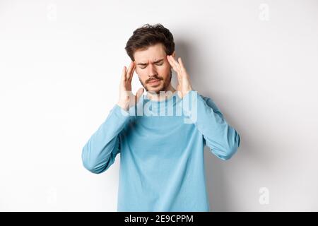 Young man having headache or hangover, touching head temples and frowning, feeling sick from migraine, standing over white background Stock Photo