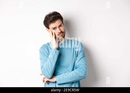 Sad young man looking frustrated, touching temple of head and frowning uneasy, standing against white background Stock Photo