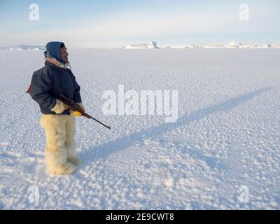 Watching a breathing hole of a seal. Inuit hunter wearing traditional trousers and boots made from polar bear fur on the sea ice of the Melville Bay n Stock Photo