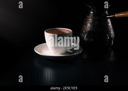 White cup of fresh hot coffee and copper cezve on black wooden table in the rays of morning light. Low key. Copy space Stock Photo