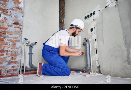 Horizontal snapshot of young plumber working with grey sewer pipes, fixing them to wall with a help of screwdriver. Side view of plumber standing on knees wearing blue uniform and white helmet Stock Photo