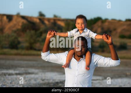 Sweet moments of fatherhood concept. Excited african american man giving piggyback ride to pretty mixed race son. Having fun on weekend spending time Stock Photo